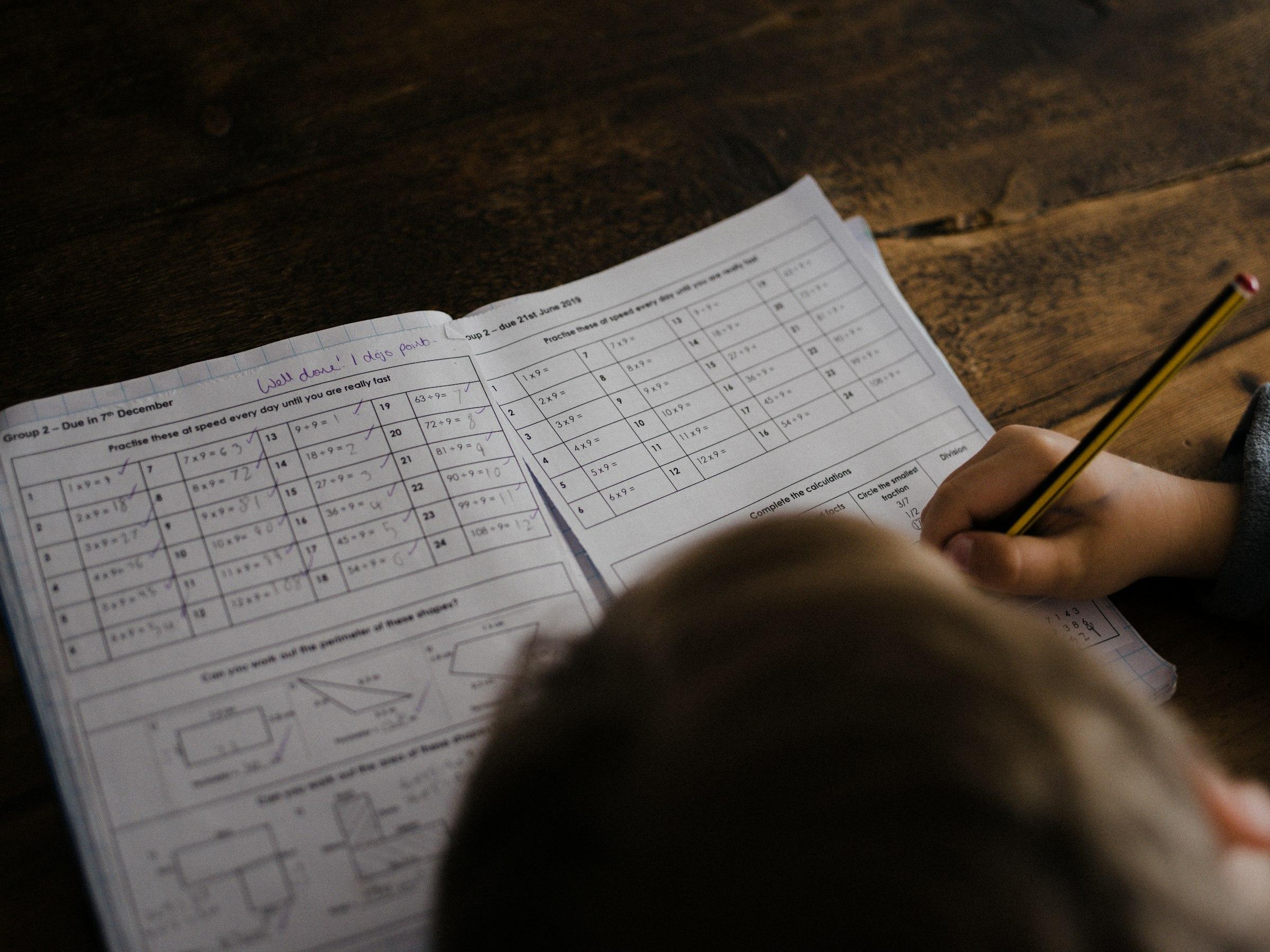 A child's hands writing out a maths problem next to a tablet